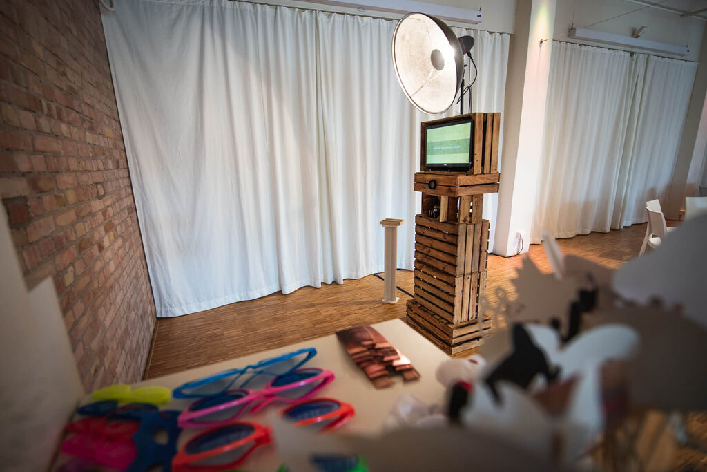 Die Holz-Fotobox mit Studioblitz aufgebaut vor einer Backsteinwand, davor Props für die Gäste