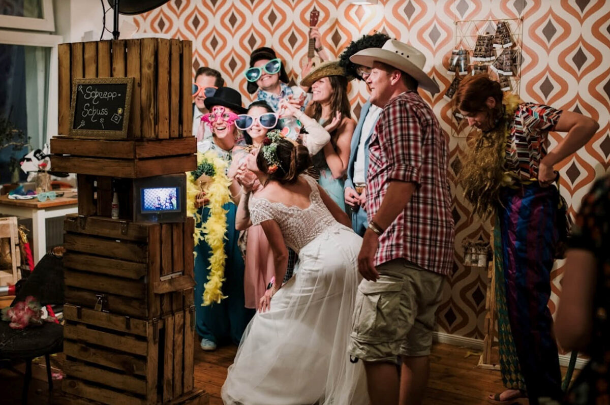 Hochzeitsgesellschaft mit Hüten, Brillen und Boas posiert lachend vor der Holz-Fotobox